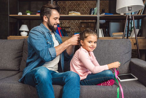 smiling father braiding his smiling daughters hair on the sofa as she plays with ribbons
