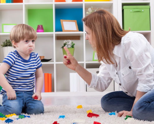 A mom scolding her son who is making a sad puppy dog face while playing with legos on the floor