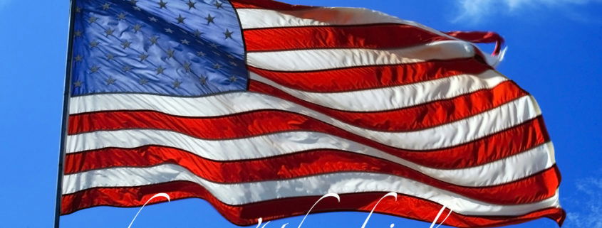 American flag waving in the wind with a bright blue sky in the background happy 4th of July written in white italics under the flag