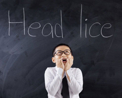 young aged school boy standing in a white collard shirt and black tie holding hands by mouth with mouth open looking at Head Lice written on the chalkboard above his head in the background