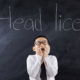 young aged school boy standing in a white collard shirt and black tie holding hands by mouth with mouth open looking at Head Lice written on the chalkboard above his head in the background