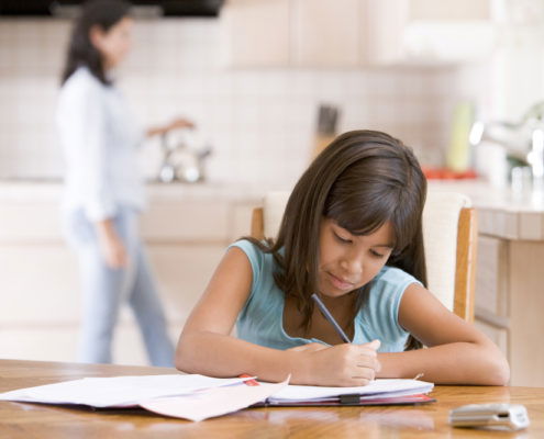 girl sitting at table doing her homework mom in the kitchen in the background putting tea on