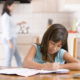 girl sitting at table doing her homework mom in the kitchen in the background putting tea on