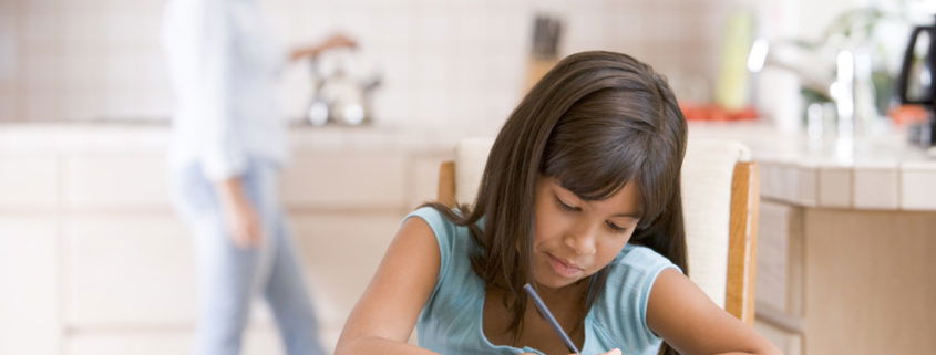 girl sitting at table doing her homework mom in the kitchen in the background putting tea on