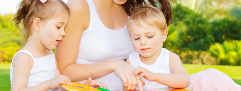 Image of cute young female with two little children read book outdoors, brother and sister with mother learning in the park in spring time, preschool education, day care, happy family concept